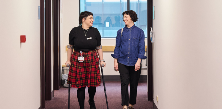 Two people walking down a hallway; one uses crutches and wears a red plaid skirt, the other wears a blue shirt and black pants.