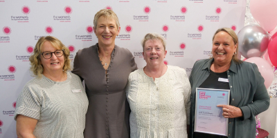A group of four smiling people in front of the Women's signage and balloons. One is holding a Staff Excellence Awards certificate.