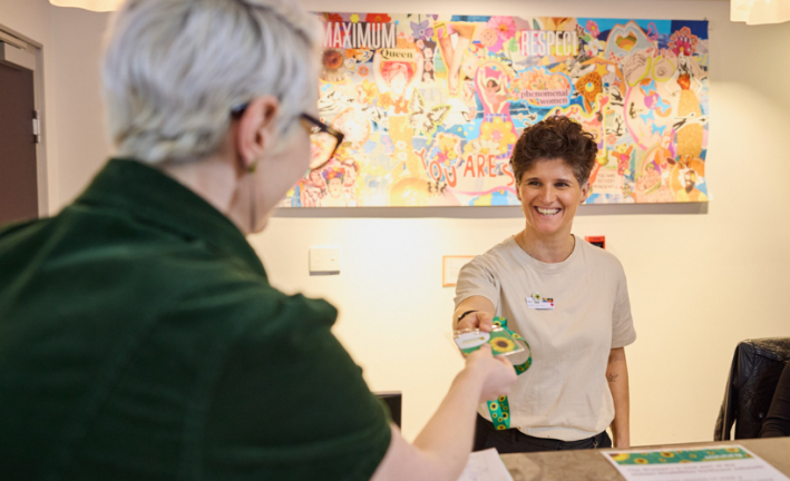 A smiling person with short hair and a beige t-shirt stands behind a counter, handing a Hidden Disability lanyard to another person with short gray hair and glasses. The background features a colorful mural with various abstract designs.